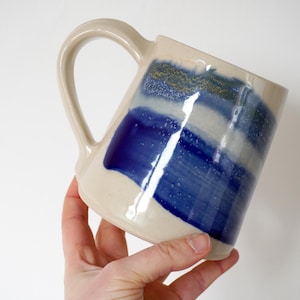 May include: Cream ceramic mug with a large handle. The mug has blue and white brushstroke designs. The mug is held at an angle, showcasing the painted details. The mug is a handmade item.