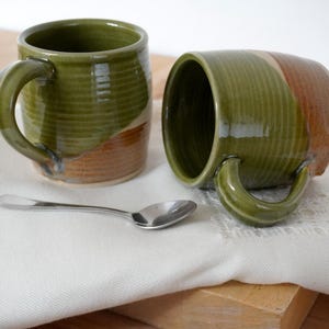 May include: Two handmade ceramic mugs with a green and brown glaze, one upright and one on its side. A small silver spoon rests on a white cloth. The mugs have a ribbed texture and curved handles, ideal for tea or coffee.