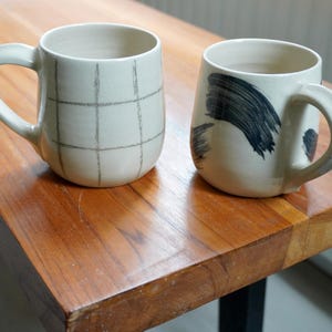 Puede incluir: Dos tazas de cerámica con asas sobre una superficie de madera. Una taza es de color blanco roto con un patrón de cuadrícula, la otra es de color blanco roto con diseños de pinceladas negras. Las tazas forman parte de una colección de utensilios de cocina.