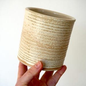 May include: A beige ceramic tumbler with a textured, horizontal striped pattern. The cylindrical cup has a slightly angled rim and is held by a hand against a white background. The pottery has a rustic, handmade appearance.