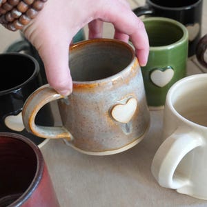 May include: A collection of ceramic mugs in various colours including black, green, cream, and brown, each featuring a small, raised heart emblem on the side. One speckled grey and brown mug is held, showing a smooth handle and a heart detail.