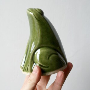 May include: A ceramic frog ornament in a glossy, olive green glaze. The frog is in a seated position with its head tilted upwards. The figurine has a smooth, rounded body and detailed features.