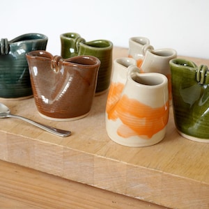 May include: A selection of handmade ceramic mugs in shades of green, brown, and cream with orange splashes. Each mug features a distinctive handle and is displayed on a wooden surface. A silver spoon is also present.