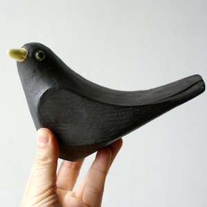 May include: A hand-carved black bird ornament with a yellow beak and green eyes. The bird has a smooth, curved body and a simple design. The figurine is held against a plain white background. A decorative item for the home.