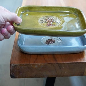 May include: Two rectangular ceramic trays, one olive green and one light blue, stacked on a wooden surface. The top tray features a tree design, while the bottom tray has a pine cone design. The trays are being held by a hand.