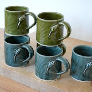 Puede incluir: Seis tazas de cerámica en tonos verdes y turquesas apiladas sobre una superficie de madera. Cada taza tiene un asa con un diseño de serpiente. Las tazas tienen una textura acanalada y forma cilíndrica.