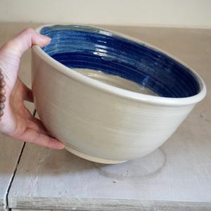 May include: A large ceramic bowl with a smooth, off-white exterior and a deep blue glazed interior. The bowl features subtle horizontal ridges along the surface, indicating a hand-thrown construction, with a wide, open circular rim.