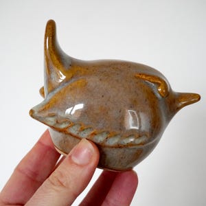 May include: A ceramic bird ornament in brown and tan hues. The bird features a curved beak and a tall, pointed crest. The ornament is held in a hand.