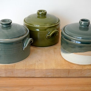 May include: Three ceramic lidded pots in shades of green sit on a wooden surface. The pots have handles and a circular design on the lids. The colours range from a deep teal to a lighter olive green.