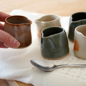 May include: A selection of small ceramic cream jugs in shades of brown, green, and off-white, with orange details. One jug is held by a hand. A silver spoon lies on a white cloth.