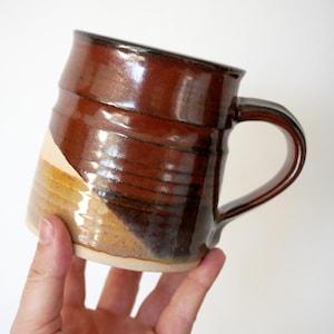 May include: A ceramic mug featuring a glossy, deep brown glaze on the upper portion and a contrasting matte, light tan finish on the lower section. The mug has a cylindrical body with a curved handle and visible horizontal ridges along the surface.