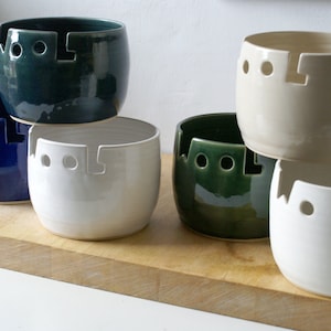 Puede incluir: Un conjunto de cuencos de cerámica para lana en varios colores, como blanco, azul, verde y crema. Cada cuenco tiene una ranura y tres agujeros para sujetar la lana. Los cuencos están apilados sobre una tabla de madera.