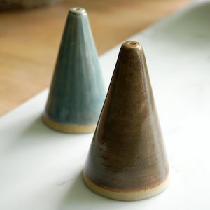 Könnte beinhalten: Zwei Keramik-Räucherstäbchenhalter in Kegelform. Einer ist in einem gedämpften Türkis, der andere in einem warmen Braun gehalten. Beide haben einen hellbraunen Sockel und ein kleines Loch oben. Die Halter stehen auf einer weißen Marmoroberfläche.