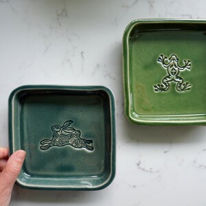 May include: Two square ceramic dishes with a glossy finish. One dish is a deep teal colour featuring an embossed rabbit design in the centre. The second dish is a vibrant grass green colour featuring an embossed frog design in the centre.