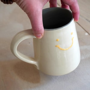May include: A cream-coloured ceramic mug with a glossy finish, featuring a simple yellow painted smiley face on the exterior. The mug has a curved handle and a dark grey interior lining, showcasing a smooth, rounded shape and a wide base.