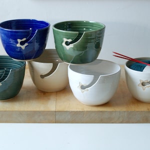 May include: A selection of ceramic yarn bowls in shades of blue, green, and white. Each bowl features a star-shaped yarn guide and a curved slot. One bowl contains yarn and knitting needles. Measurements are in cm.