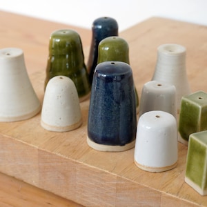 May include: A selection of ceramic salt and pepper shakers in diverse shapes and colours. The set features white, green, and blue shakers, some cylindrical and others square. The shakers are displayed on a wooden chopping board.