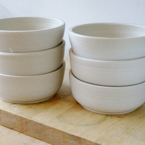 May include: A set of six white ceramic bowls stacked in two piles of three. The bowls have a slightly textured surface and are a simple, elegant design.