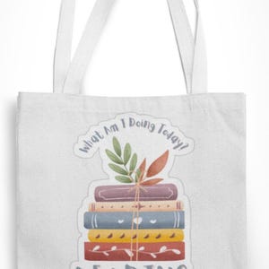 May include: White tote bag featuring a graphic of a stack of books tied with twine, adorned with leaves. The text "What Am I Doing Today?" curves above the books, with "READING" printed below.