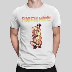 May include: White t-shirt with a pixelated graphic of two figures in a fighting pose, and the text "FINISH HIM!!" in a yellow and red gradient font. A casual wear item.