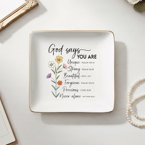 Puede incluir: Una bandeja cuadrada de cerámica blanca con ribete dorado. La bandeja presenta el texto "Dios dice que eres" con un diseño floral y palabras inspiradoras. Un espejo con marco dorado, un calendario y un collar de perlas también están en la imagen.