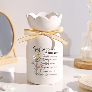 Puede incluir: Un jarrón de cerámica blanca con una parte superior festoneada y una cinta dorada atada alrededor del cuello. El jarrón tiene el texto "God says YOU ARE" con acentos florales y palabras inspiradoras. Un artículo decorativo.
