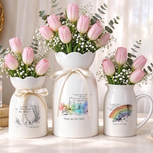 May include: Three white ceramic vases and a pitcher filled with pink tulips and baby's breath. Each vessel features Winnie the Pooh illustrations and text. The centre vase is tied with a cream ribbon. The text includes quotes and personalised names.