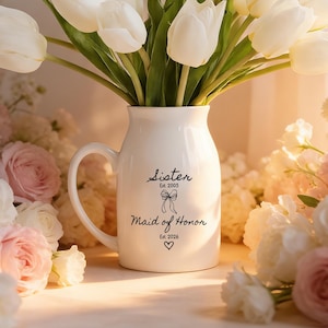 May include: A white ceramic vase with a handle, filled with white tulips. The vase has the words "Sister Est. 2005" and "Maid of Honor Est. 2026" printed on it. The vase is surrounded by pink and white roses.