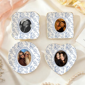 Puede incluir: Cuatro portafotos decorativos de cerámica en forma cuadrada, redonda y de corazón. Cada soporte presenta un estampado floral azul sobre fondo blanco y un borde dorado. Los soportes contienen fotografías en blanco y negro o en color.