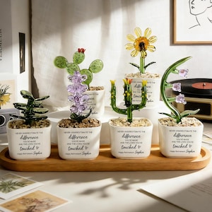 Può includere: Una collezione di sculture di piante in vetro in piccoli vasi bianchi, disposte su un vassoio di legno. Le sculture includono un cactus, lavanda, girasole e altri disegni floreali. Ogni vaso ha il testo "NEVER UNDERESTIMATE THE DIFFERENCE YOU'VE MADE AND THE LIVES YOU'VE touched Happy Retirement Sophia."