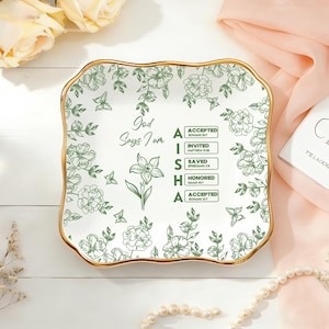 Puede incluir: Un plato decorativo cuadrado de cerámica blanca con borde dorado y diseño floral verde. El plato tiene el texto "Dios dice que soy" y el nombre "AISHA" con versículos bíblicos asociados. El plato está rodeado de flores y un collar de perlas.