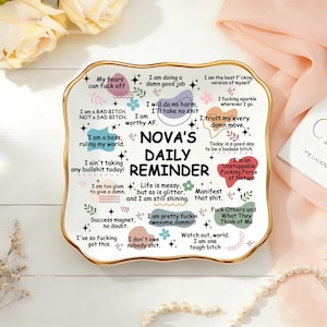 Könnte beinhalten: Ein weißes Keramik-Tageserinnerungs-Tablett mit goldenem Rand, das verschiedene motivierende Sätze und den Text "NOVA'S DAILY REMINDER" enthält. Das Tablett ist mit bunten Blumen- und Sternakzenten verziert.