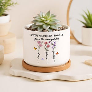 Puede incluir: Maceta de cerámica blanca con una suculenta, con el texto "Sisters are different flowers from the same garden". La maceta tiene un platillo de madera e incluye ilustraciones florales y nombres.