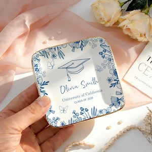 Może przedstawiać: Kwadratowy ceramiczny talerz pamiątkowy z okazji ukończenia studiów ze złotą obwódką. Biały talerz zdobią niebieskie wzory kwiatowe i motyle, czapka absolwenta oraz napis "Olivia Smith, University of California, CLASS OF 2026."