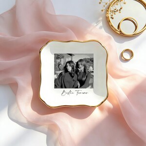 May include: A square white ceramic trinket dish with scalloped edges and a gold-toned rim. The center features a black and white photograph of two people with the cursive text Bestie Forever printed underneath the image on the white surface.