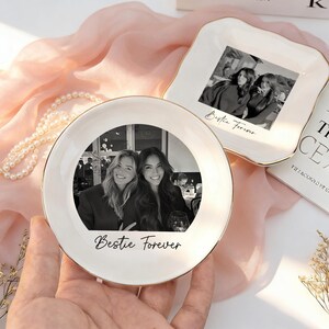 May include: Two white ceramic trinket dishes with gold-toned rims. One dish is round and the other is square with scalloped edges. Both feature a black and white photograph of two people and the cursive text "Bestie Forever" printed on them.