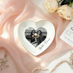 May include: A white heart-shaped ceramic trinket dish with a gold-toned rim. The center features a black and white photograph of two people, with a gold ring with a white pearl resting on top. The text "Bestie Forever" is printed in black script.