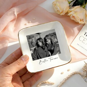 May include: A square white ceramic trinket dish with a gold-toned rim, featuring a black and white photograph of two people and the printed text "Bestie Forever" in a cursive script centered on the surface of the dish.