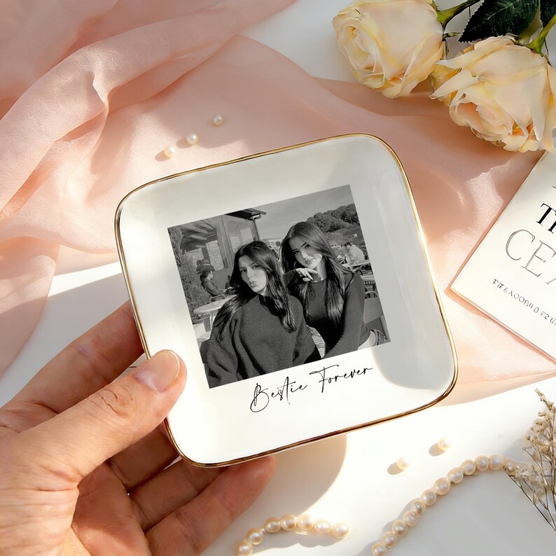May include: A square white ceramic trinket dish with a gold-toned rim, featuring a black and white photograph of two people and the printed text "Bestie Forever" in a cursive script centered on the surface of the dish.