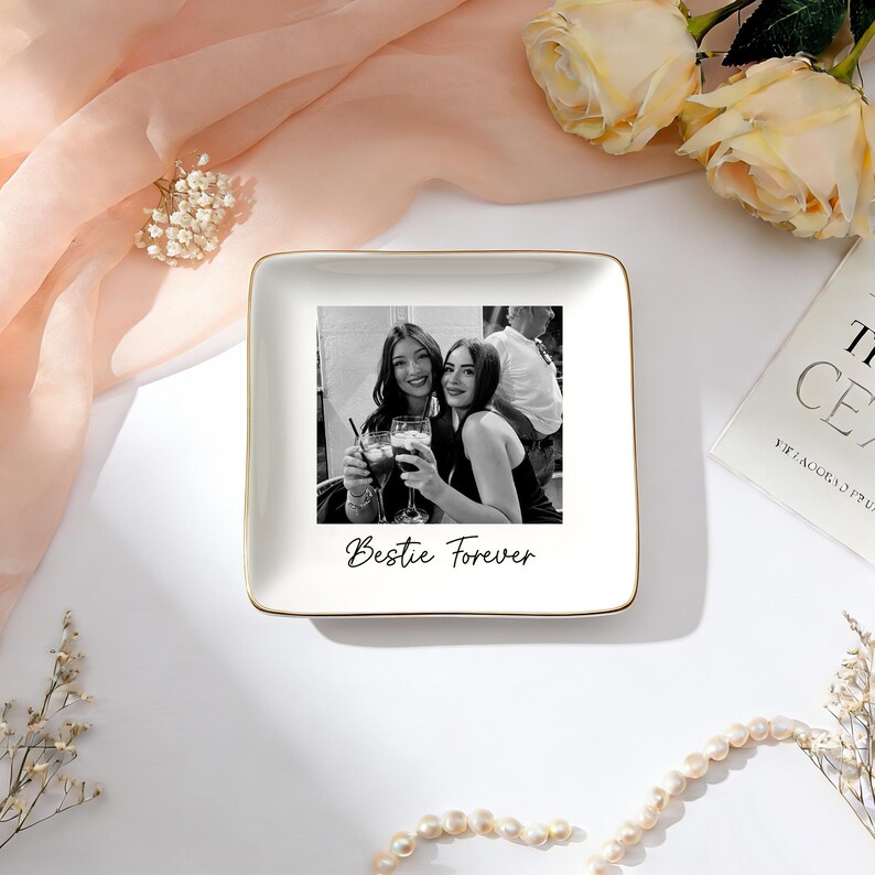 May include: A square white ceramic trinket dish with a gold-toned rim. The center features a black and white photograph of two people holding glasses, with the black printed text "Bestie Forever" positioned directly below the photograph.