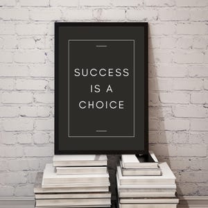 May include: A framed black print with the words "SUCCESS IS A CHOICE" in white. The print is displayed on a stack of books against a white brick wall. The frame has a simple, modern design.