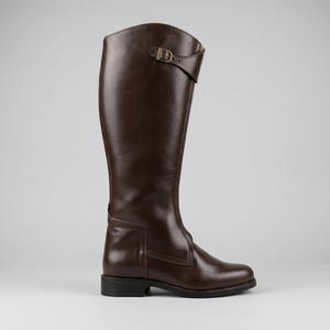 May include: A tall, brown leather riding boot. The boot features a buckle detail at the top and a small, rectangular accent near the ankle. The boot has a flat sole and a smooth, polished finish, suitable for equestrian activities.