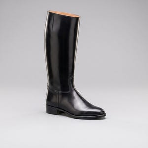 Puede incluir: Una bota de montar alta de cuero negro con un acabado liso y pulido. La bota presenta una caña estructurada, una punta redondeada, un tacón bajo y una costura visible que recorre verticalmente el lateral de la caña, con forro interior color canela.