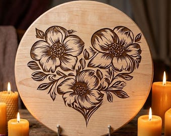 May include: A round, light-colored wooden plaque with a floral heart design. The flowers and leaves are intricately carved in a darker brown. The plaque is supported by two metal stands. The background includes lit candles and a key.