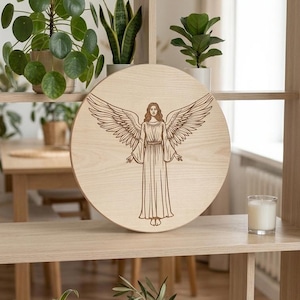 Op de afbeelding: Ronde houten plaat met een gegraveerde afbeelding van een engel met uitgespreide vleugels. De engel draagt een lange jurk. De lichtgekleurde plaat staat op een houten plank, met planten op de achtergrond.