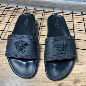 May include: A pair of black rubber slides featuring a wide strap with an embossed Medusa head logo and a Greek key pattern. The footbed is molded with the text Versace Made in Italy printed in white on the heel area of each sandal.