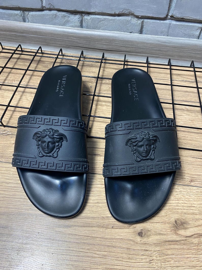 May include: A pair of black rubber slides featuring a wide strap with an embossed Medusa head logo and a Greek key pattern. The footbed is molded with the text Versace Made in Italy printed in white on the heel area of each sandal.