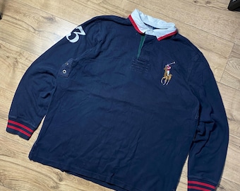 May include: A navy blue long-sleeve rugby shirt featuring a white collar with red trim, a button placket, and a large embroidered polo player logo on the chest. The right sleeve displays a white number 3, and the cuffs have red and navy stripes.