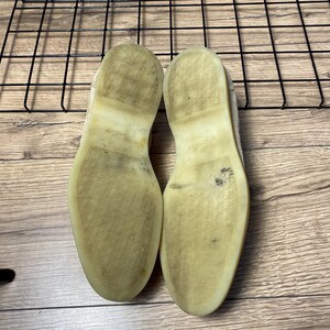 Op de afbeelding: Een paar schoenen met lichtgekleurde, doorschijnende rubberen zolen. De zolen zijn plat en vertonen gebruikssporen met donkere vegen en vuil. De textuur van de zolen is glad met een subtiel in reli&euml;f aangebracht logo in het midden.