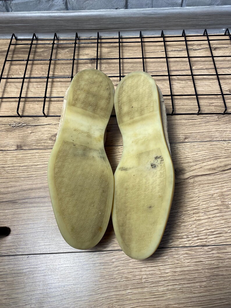 Op de afbeelding: Een paar schoenen met lichtgekleurde, doorschijnende rubberen zolen. De zolen zijn plat en vertonen gebruikssporen met donkere vegen en vuil. De textuur van de zolen is glad met een subtiel in reli&euml;f aangebracht logo in het midden.